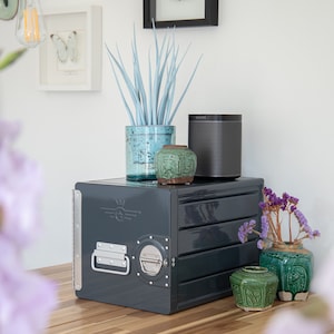 May include: A dark gray metal storage box with a handle and a SONOS speaker on top. The box is sitting on a wooden surface with a potted plant, a green vase with purple flowers, and two green ceramic jars.