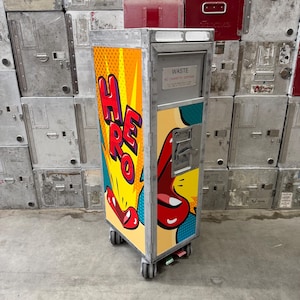 May include: A tall, rectangular metal airline service cart featuring a pop art graphic with the word HERO in large red letters, yellow sunburst patterns, and red lips. The cart has a silver frame, four wheels, and a metal plate labelled WASTE.