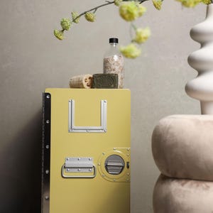 May include: A yellow storage cabinet with metal accents, a handle, and wheels. The cabinet has a rectangular shape with a U-shaped metal detail. A bottle, soap, and loofah sit on top. A vase with flowers is in the background.