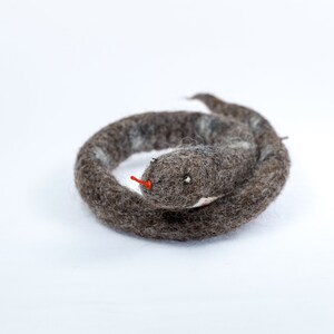 May include: A grey and white felt snake toy curled up in a circle. The snake has a red felt tongue and black eyes.