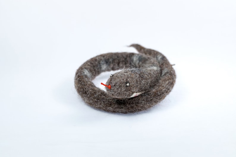 May include: A grey and white felt snake toy curled up in a circle. The snake has a red felt tongue and black eyes.