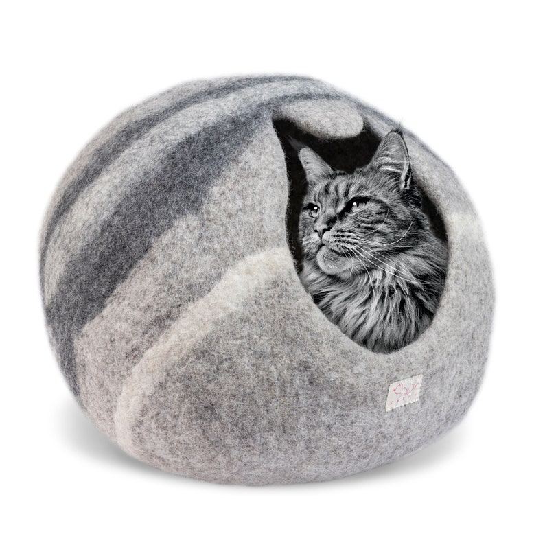 May include: A gray and white felt cat bed shaped like a sphere with a cat inside. The cat bed has a hole for the cat to enter and exit. The cat is looking out of the hole.