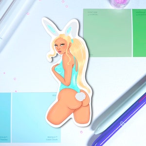 May include: A sticker featuring a blonde woman in a light blue bunny costume. The sticker is on a white surface with paint swatches, pens, and pink star-shaped confetti. The paint swatches are shades of blue and green.