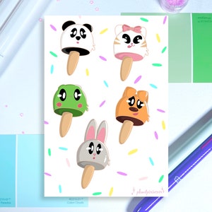 Könnte beinhalten: Ein Druck mit fünf Cartoon-Eis am Stiel in Form von Tieren: einem Panda, einer Katze, einem Frosch, einem Hund und einem Hasen. Jedes Eis am Stiel hat einen Holzstab und ist mit bunten Streuseln verziert. Der Hintergrund ist weiß.
