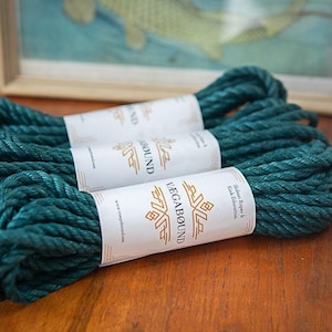 May include: Three coils of dark teal twisted rope, each secured with a white paper band featuring the gold text VAGABOUND and the words Shibari Ropes & Kink Education. The rope has a thick, textured, multi-strand construction.