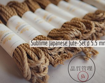 Op de afbeelding: Een set opgerolde bruine jutekoorden, elk omwikkeld met een wit papieren label met gouden tekst en de merknaam VAGABOUND. De tekst Sublime Japanese Jute-Set ø 5.5 mm is zichtbaar, samen met een cirkelvormige stempel met Japanse karakters.