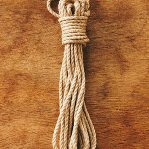 May include: A length of natural tan jute rope coiled into a long, vertical bundle. The rope features a twisted fibre construction with a textured surface, secured at the top with several tight wraps of the same material to maintain its shape.