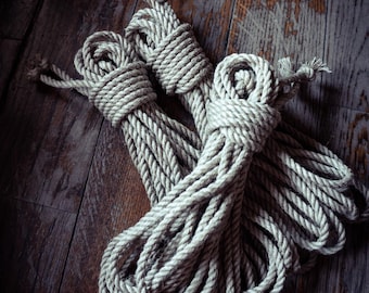 May include: Two coils of thick, natural-coloured jute rope, each neatly wound and secured with a central binding knot. The ropes feature a twisted fibre construction with a textured surface, showing visible strands and frayed ends at the top.
