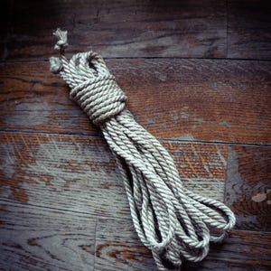 May include: A coiled length of thick, natural-colored jute rope featuring a tightly twisted fiber construction. The rope is bundled with a secure knot at the top, with several loose loops extending downward, showcasing the textured, fibrous surface.
