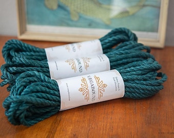 May include: Three coils of dark teal twisted rope, each secured with a white paper band featuring the gold text VAGABOUND and the phrase Shibari Ropes & Kink Education, along with a website URL printed on the side of the paper labels.