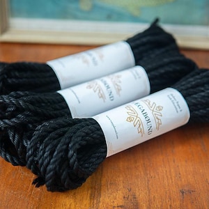 May include: Three coils of black twisted rope, each secured with a white paper band. The bands feature the text "MEGABOUND" in a bold font, accompanied by a geometric logo and the phrase "Jute Rope & Silk Education" printed in black ink.