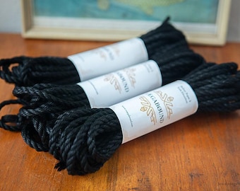 May include: Three coils of black twisted rope, each secured with a white paper band. The bands feature the text "MEGABOUND" in a bold font, accompanied by a geometric logo and the phrase "Jute Rope & Silk Education" printed in black ink.