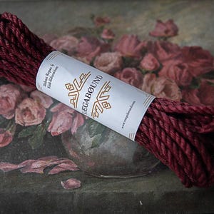 Puede incluir: Un rollo de cuerda trenzada de color burdeos oscuro asegurado con una banda de papel blanca. La banda presenta el texto dorado VAGABØUND, la frase Shibari Ropes & Kink Education y la dirección web www.ropegabound.com en tinta negra.