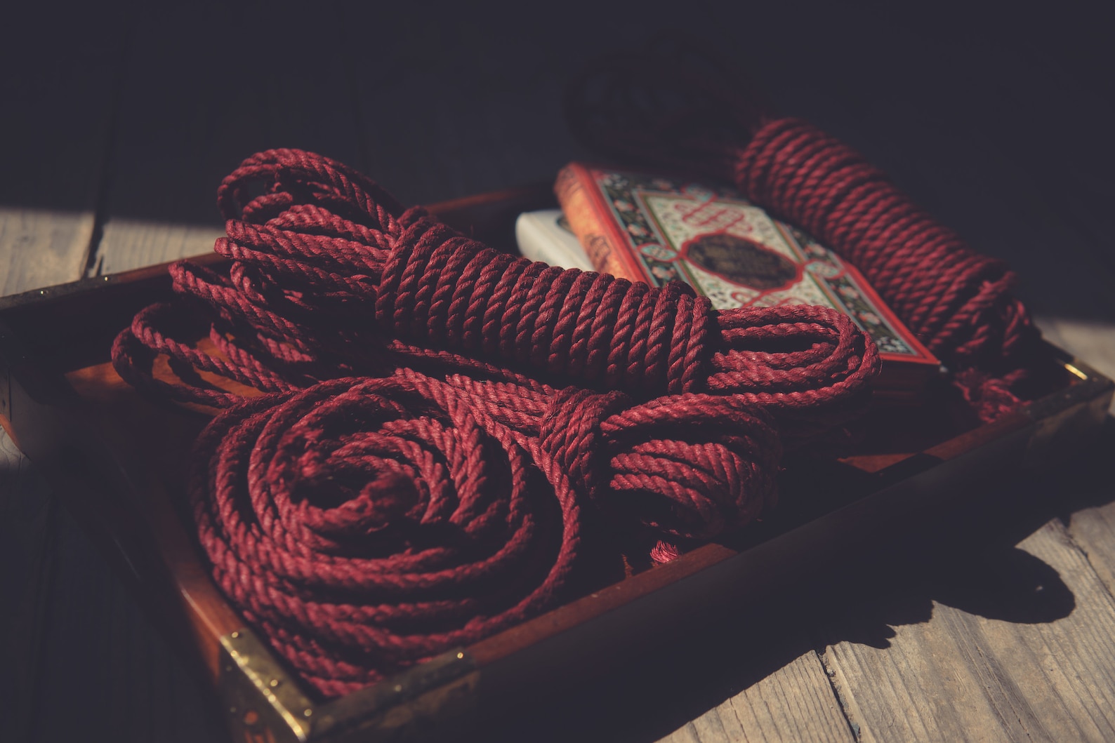Deep red hemp bondage ropes coiled on a wooden tray beside a decorative box, in warm light, ready for shibari or kinbaku tying.