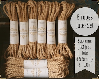 Op de afbeelding: Een set van acht beige jute touwen, elk omwikkeld met een witte papieren band met het label VÆGABØUND. De tekst rechts luidt 8 ropes Jute-Set, Supreme JBO free Jute, 5,5 mm diameter en 8-10 m lengte. De touwen zijn gestapeld.
