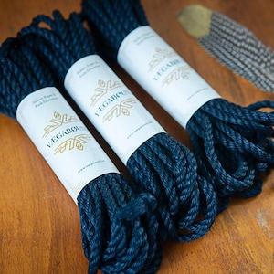 May include: Three coils of dark navy blue twisted rope, each secured with a white paper band featuring the gold logo and text VÆGABØUND. A single brown and white patterned feather lies to the right of the rope coils.