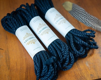 May include: Three coils of dark navy blue twisted rope, each secured with a white paper band featuring the gold logo and text VÆGABØUND. A single brown and white patterned feather lies to the right of the rope coils.
