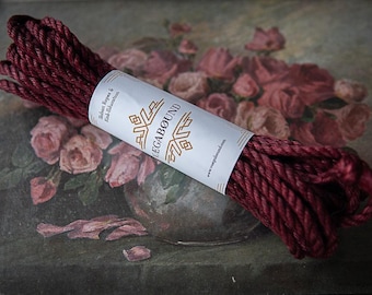 May include: A coil of deep burgundy twisted rope secured with a white paper band. The band features the text "VEGABØUND" in black, along with the smaller text "Shibari Ropes & Kink-Education" and the website "www.vegabound.com" printed in black.