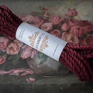 Può includere: Una matassa di corda ritorta color bordeaux scuro fissata con una fascia di carta bianca. La fascia riporta il testo VAGABOUND in un carattere stilizzato, insieme alle parole Shibari Ropes & Kink-Education e l'indirizzo web www.vagabound.com.