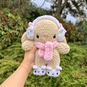May include: A plush, beige bunny wearing white earmuffs with pink accents, a pink scarf, and white booties. The bunny has black eyes and a pink nose. The bunny is held in a person's hand against a backdrop of green foliage.