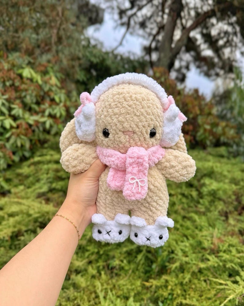 May include: A plush, beige bunny wearing white earmuffs with pink accents, a pink scarf, and white booties. The bunny has black eyes and a pink nose. The bunny is held in a person's hand against a backdrop of green foliage.
