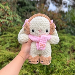 May include: A plush, cream-colored bunny with brown earmuffs, feet, and a pink scarf. The bunny has black eyes and is made of a soft, textured material. The scarf has a bow detail. The bunny is a handmade item.