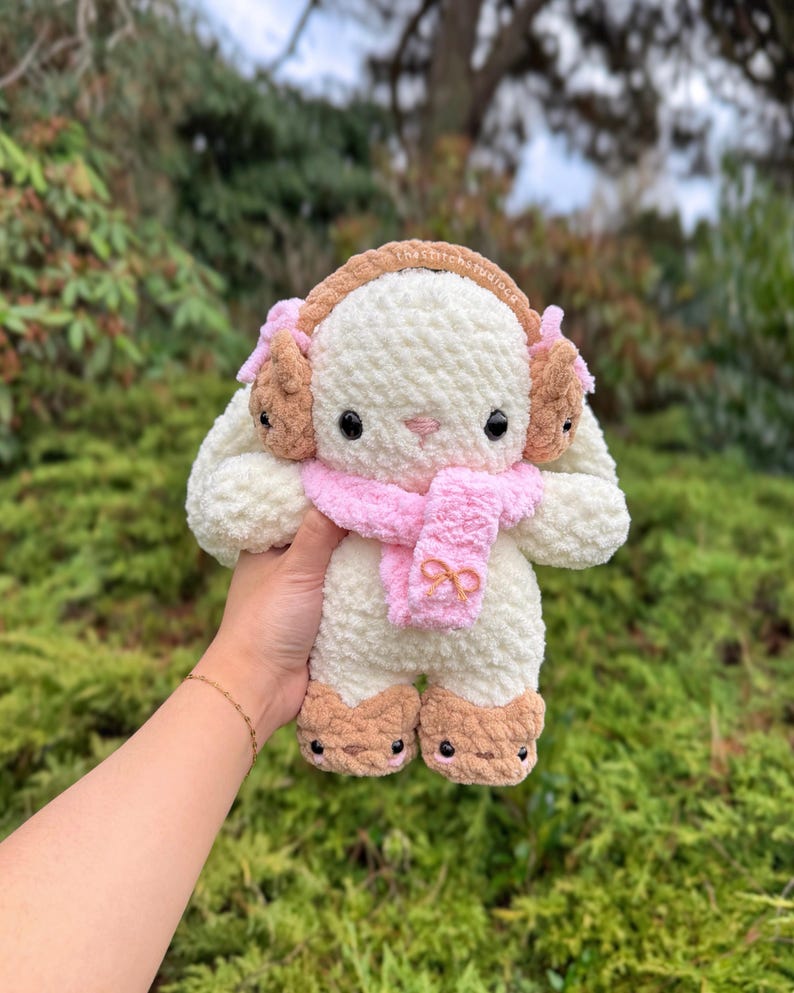 May include: A plush, cream-colored bunny with brown earmuffs, feet, and a pink scarf. The bunny has black eyes and is made of a soft, textured material. The scarf has a bow detail. The bunny is a handmade item.