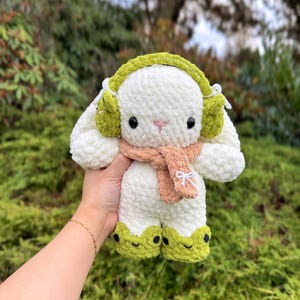 May include: A handmade, plush white bunny with green earmuffs, feet, and a tan scarf. The bunny has black eyes and a pink nose. The scarf is tied with a white bow. The bunny is a soft, cuddly toy.