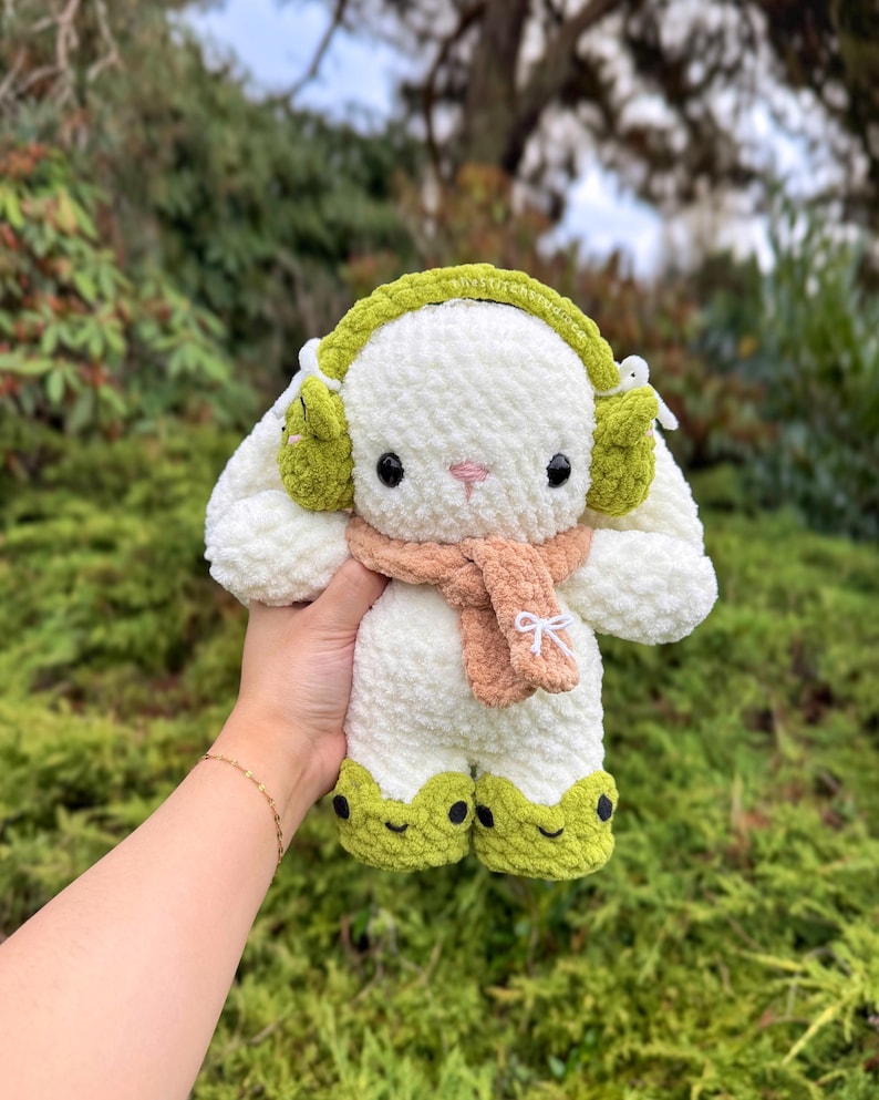 May include: A handmade, plush white bunny with green earmuffs, feet, and a tan scarf. The bunny has black eyes and a pink nose. The scarf is tied with a white bow. The bunny is a soft, cuddly toy.