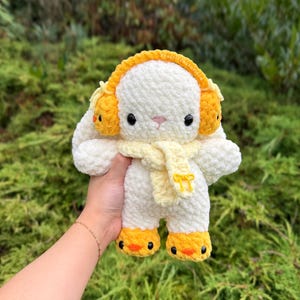 May include: A handmade, white crochet bunny plushie with yellow earmuffs, a yellow scarf, and yellow feet. The bunny has black eyes and a pink nose. The plushie is held in front of a green, leafy background.
