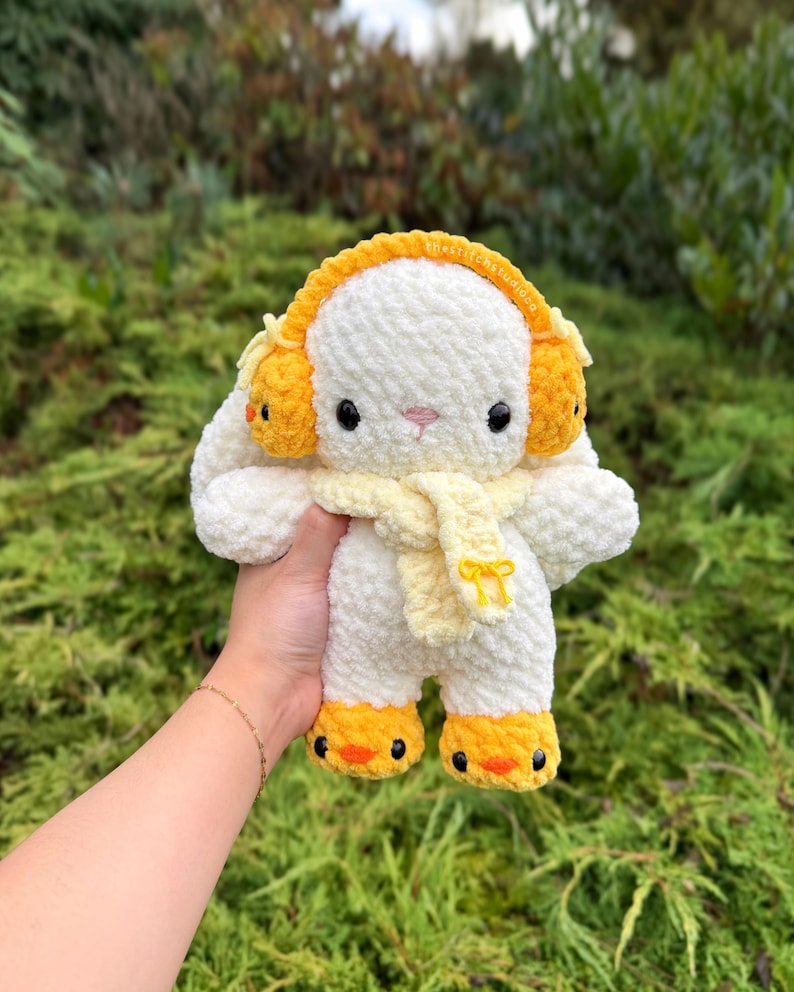 May include: A handmade, white crochet bunny plushie with yellow earmuffs, a yellow scarf, and yellow feet. The bunny has black eyes and a pink nose. The plushie is held in front of a green, leafy background.
