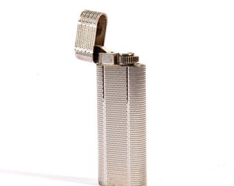 May include: A silver-colored lighter with a textured, geometric pattern. The top is open, revealing the inner mechanism. The lighter has a rectangular shape with rounded edges and a small, circular wheel for ignition.
