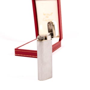 Puede incluir: Un encendedor plateado de forma rectangular, abierto para revelar un interior blanco y adornos dorados. El encendedor se muestra en una caja roja con interior blanco y letras doradas.