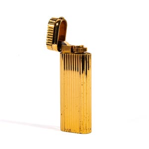 May include: A gold-coloured lighter with a ribbed design. The top is open, revealing the inner mechanism. The lighter has a rectangular shape with rounded edges and a textured surface. It is set against a white background.