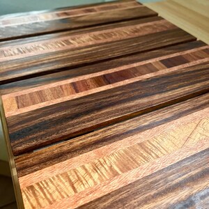 May include: A wooden cutting board with a striped design. The board features alternating strips of dark brown and light brown wood, creating a visually appealing pattern. The wood grain is visible, adding texture and warmth to the design.