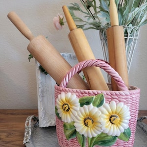May include: A pink ceramic basket with a woven texture and daisy embellishments holds several wooden rolling pins. The basket has a braided handle and sits on a silver tray, with flowers in the background.