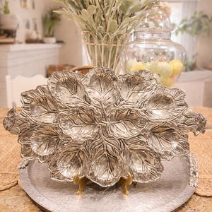 May include: A silver-colored, leaf-shaped serving dish with intricate veining details, resting on a hammered metal tray. The dish is supported by small, gold-colored feet. The background includes a vase of peach-colored flowers and a glass jar.