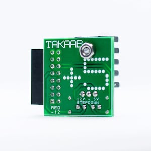 May include: A green circuit board with the text "TAKAAE" and "12V - 5V STEP DOWN" printed on it. The board has a black connector on the left side and a row of pins on the right side. The text "RED -12" is printed on the bottom left corner of the board.