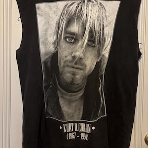 May include: Black sleeveless t-shirt with a black and white portrait of Kurt Cobain. The image includes the text "KURT D. COBAIN (1967-1994)" in white. The shirt has distressed edges.