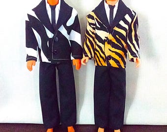 May include: Two dolls wearing black pants and patterned jackets. One jacket has a black and white zebra print, the other has a brown and white tiger print.