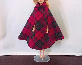 May include: A doll wearing a red and black plaid cape with a hood. The cape features a classic tartan pattern with intersecting lines and is fastened at the neck. The doll is supported by a clear plastic stand that holds it in an upright position.