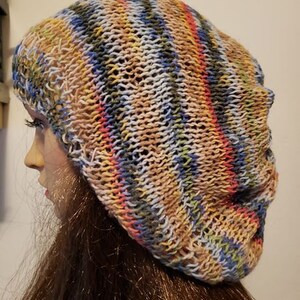 Könnte beinhalten: Eine mehrfarbige gestreifte Strickmütze mit einem lockeren Sitz. Die Mütze ist aus einem weichen, leichten Garn gefertigt und hat eine Vielzahl von Farben, darunter Blau, Grün, Pink und Beige.