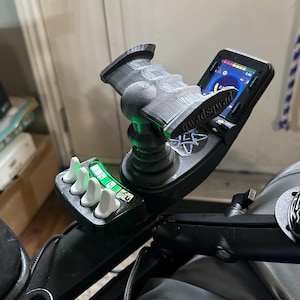 May include: A black and grey joystick controller with a screen showing a user interface. The controller features a textured grip and several white buttons. The text "@quadsqwad" is visible on the device.