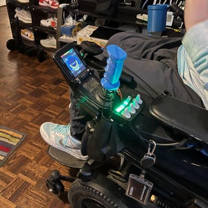 Puede incluir: Una silla de ruedas eléctrica negra con un joystick azul y una pantalla digital. La silla de ruedas tiene un asiento y reposabrazos negros. Las ruedas son negras con un borde plateado. La persona lleva zapatillas deportivas azules y blancas.