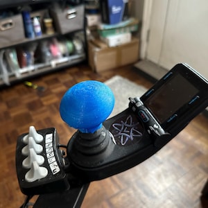 Puede incluir: Un joystick azul con una bola azul en la parte superior. El joystick tiene una base negra y un ribete plateado. Hay cuatro botones blancos en el lateral del joystick. El joystick está en un suelo de madera.