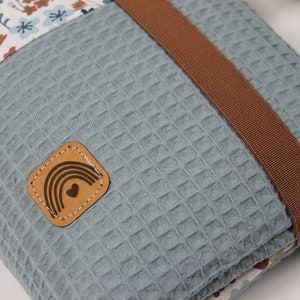May include: A blue waffle-textured pouch with a brown strap and a small leather patch with a rainbow and heart design. The pouch has a floral pattern on one side.