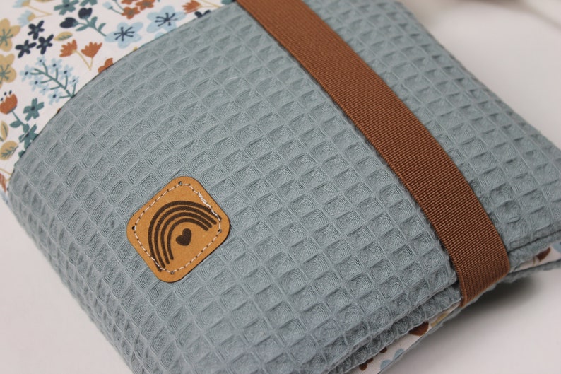 May include: A blue waffle-textured pouch with a brown strap and a small leather patch with a rainbow and heart design. The pouch has a floral pattern on one side.