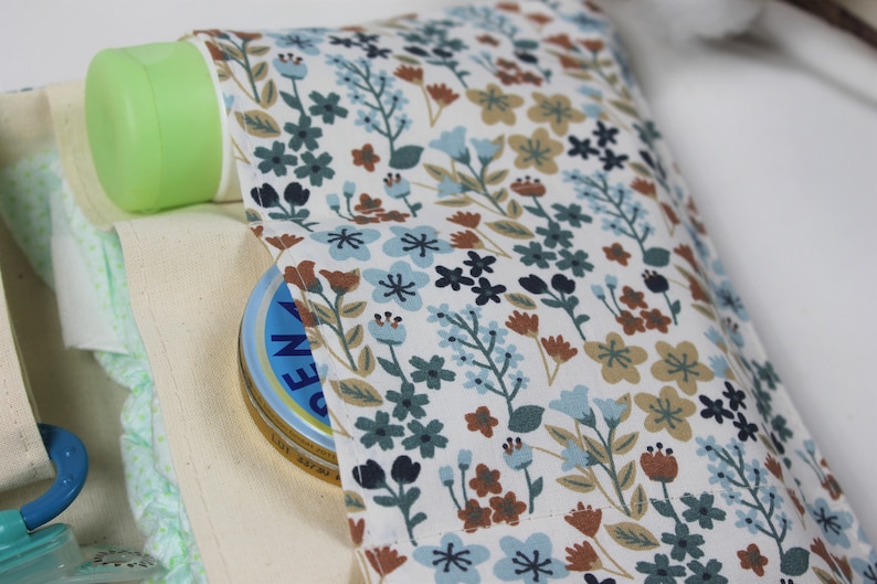 May include: A white canvas bag with a floral print lining. The bag is filled with baby supplies, including a green bottle, a blue pacifier, and a white jar of cream.