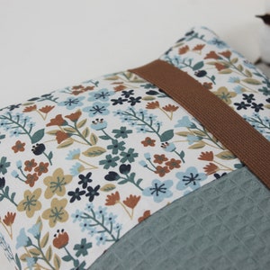 May include: A small, rectangular pillow with a floral pattern in shades of blue, brown, and white. The pillow has a brown strap and a textured, teal fabric on the bottom.