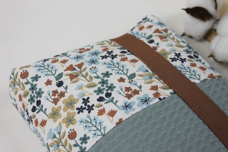 May include: A small, rectangular pillow with a floral pattern in shades of blue, brown, and white. The pillow has a brown strap and a textured, teal fabric on the bottom.